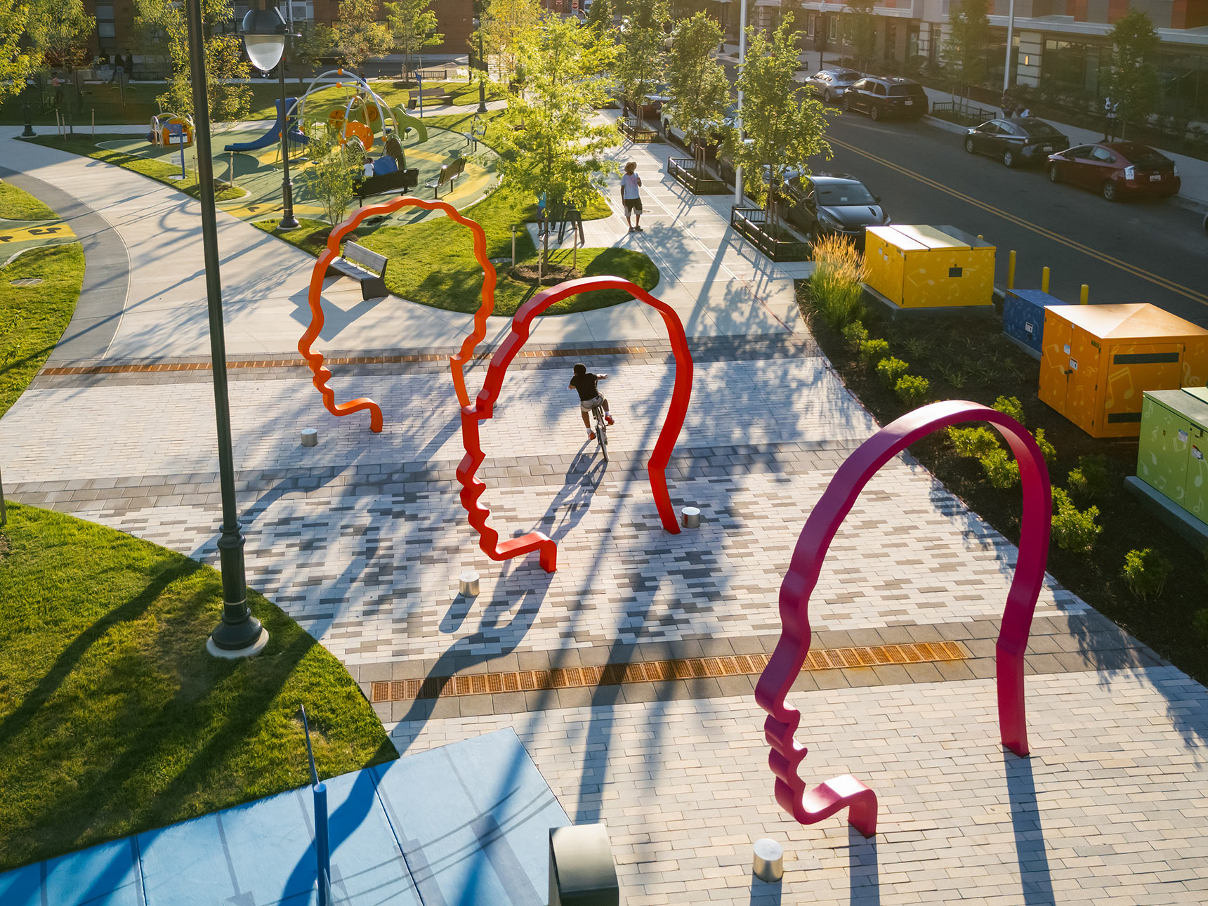 A kid rides a bicycle through three monumental 12-foot-tall silhouette portraits in a colorful gradient of orange, red, and purple.