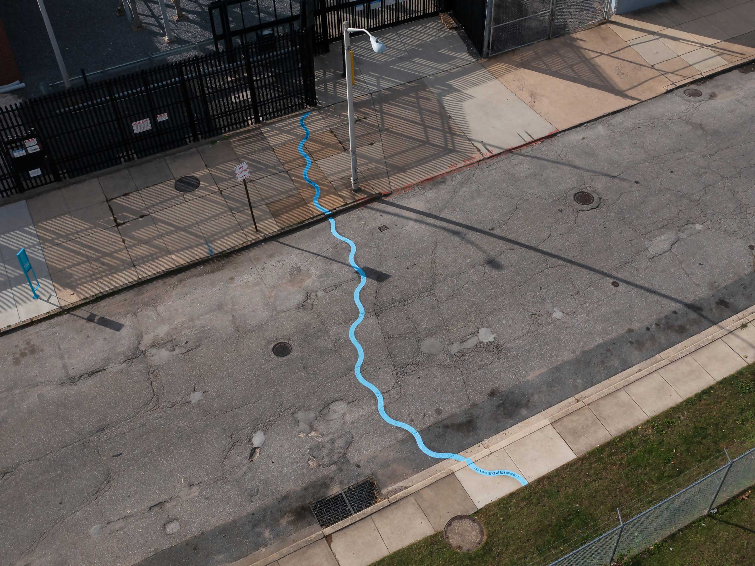 Aerial photo of a public art installation mapping the path of an underground stream. A bright blue wavy line snakes all the ways across a roadway and sidewalks between a grass strip and a black metal fence.