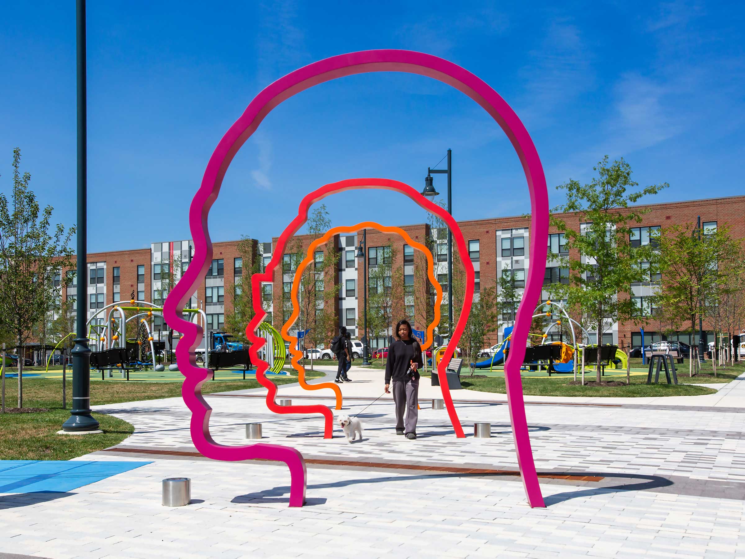 A woman walks a dog under three 12-foot-tall silhouette portrait archways in a colorful gradient of orange, red, and purple.