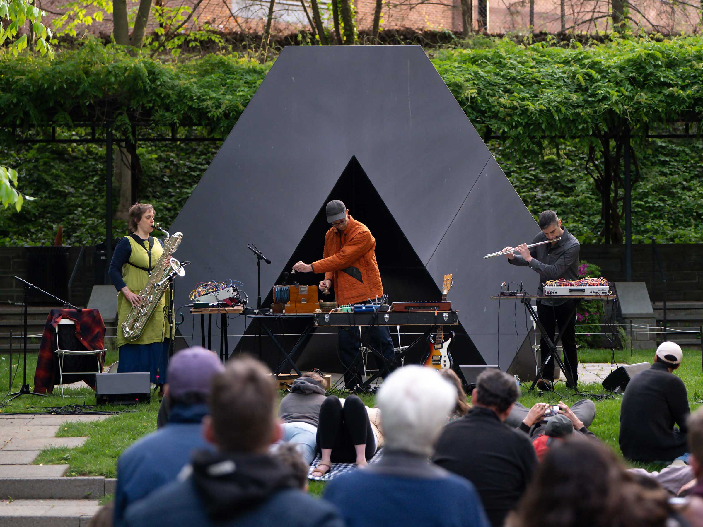 Musicians Bruce Willen, Jess Keyes, and Jason Charney performing Music for a Ghost River in the BMA Sculpture Garden in front of a black triangular Tony Smith sculpture. Dozens of people lay on the grass listening.