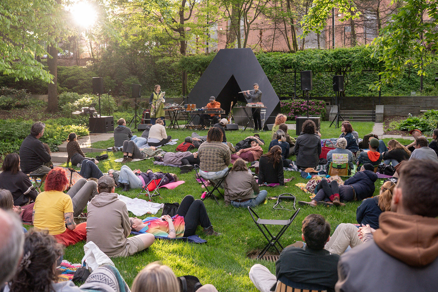 Musicians Bruce Willen, Jess Keyes, and Jason Charney performing Music for a Ghost River in the BMA Sculpture Garden in front of a black triangular Tony Smith sculpture. Dozens of people lay on the grass listening.