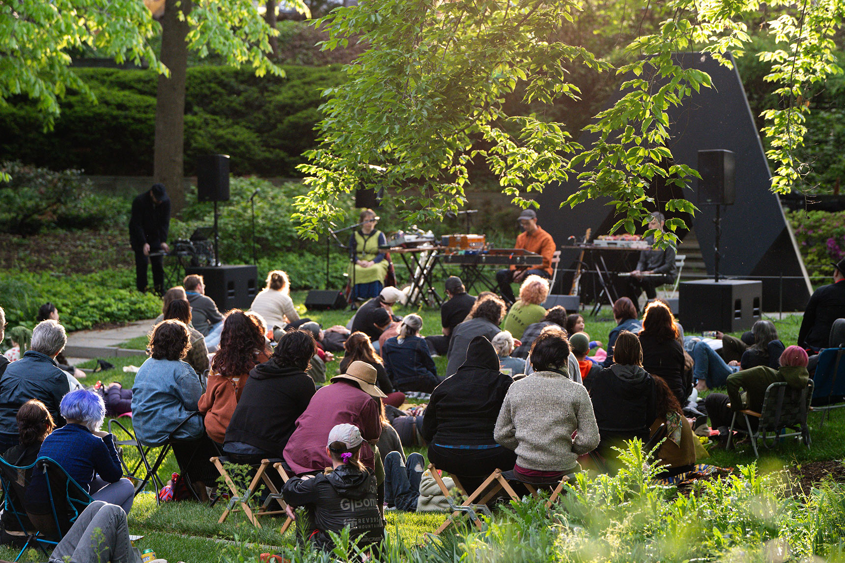 Musicians Bruce Willen, Jess Keyes, and Jason Charney performing Music for a Ghost River in the BMA Sculpture Garden in front of a black triangular Tony Smith sculpture. Dozens of people lay on the grass listening quietly