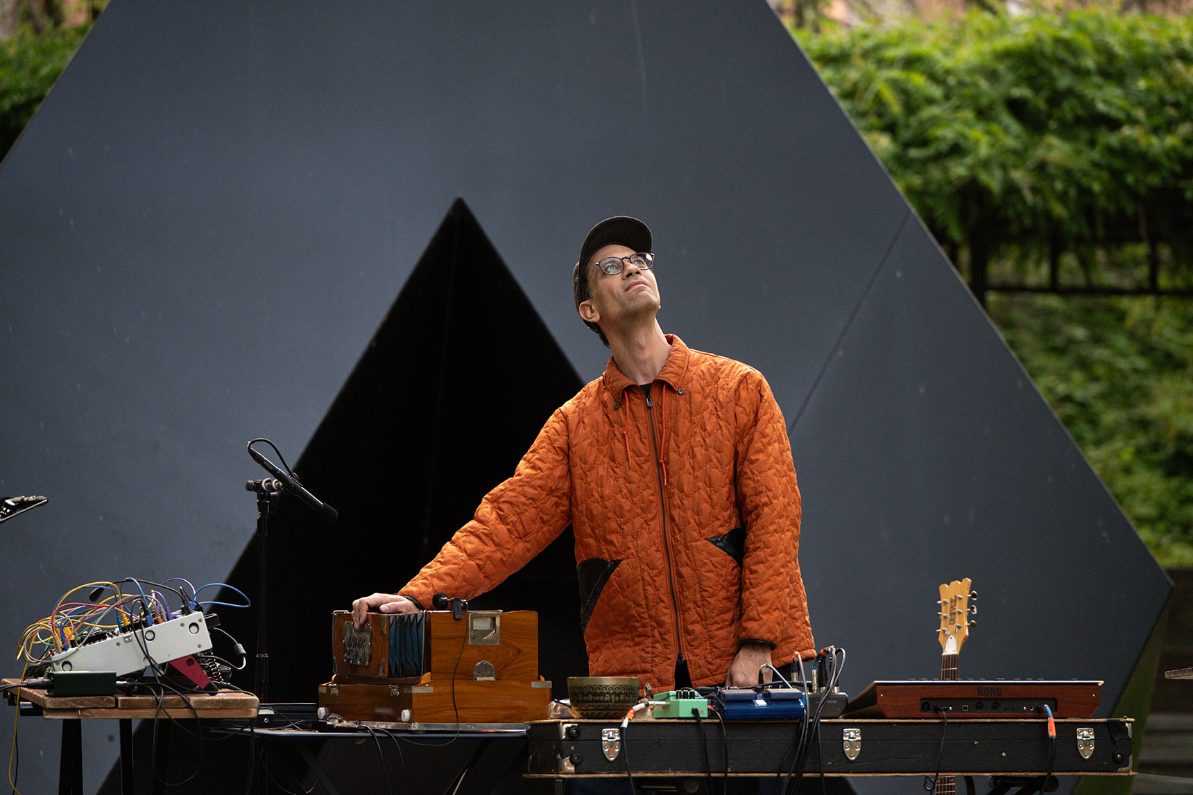 Bruce Willen playing harmonium, looking up at trees in the BMA Sculpture Garden in front of a black triangular Tony Smith sculpture